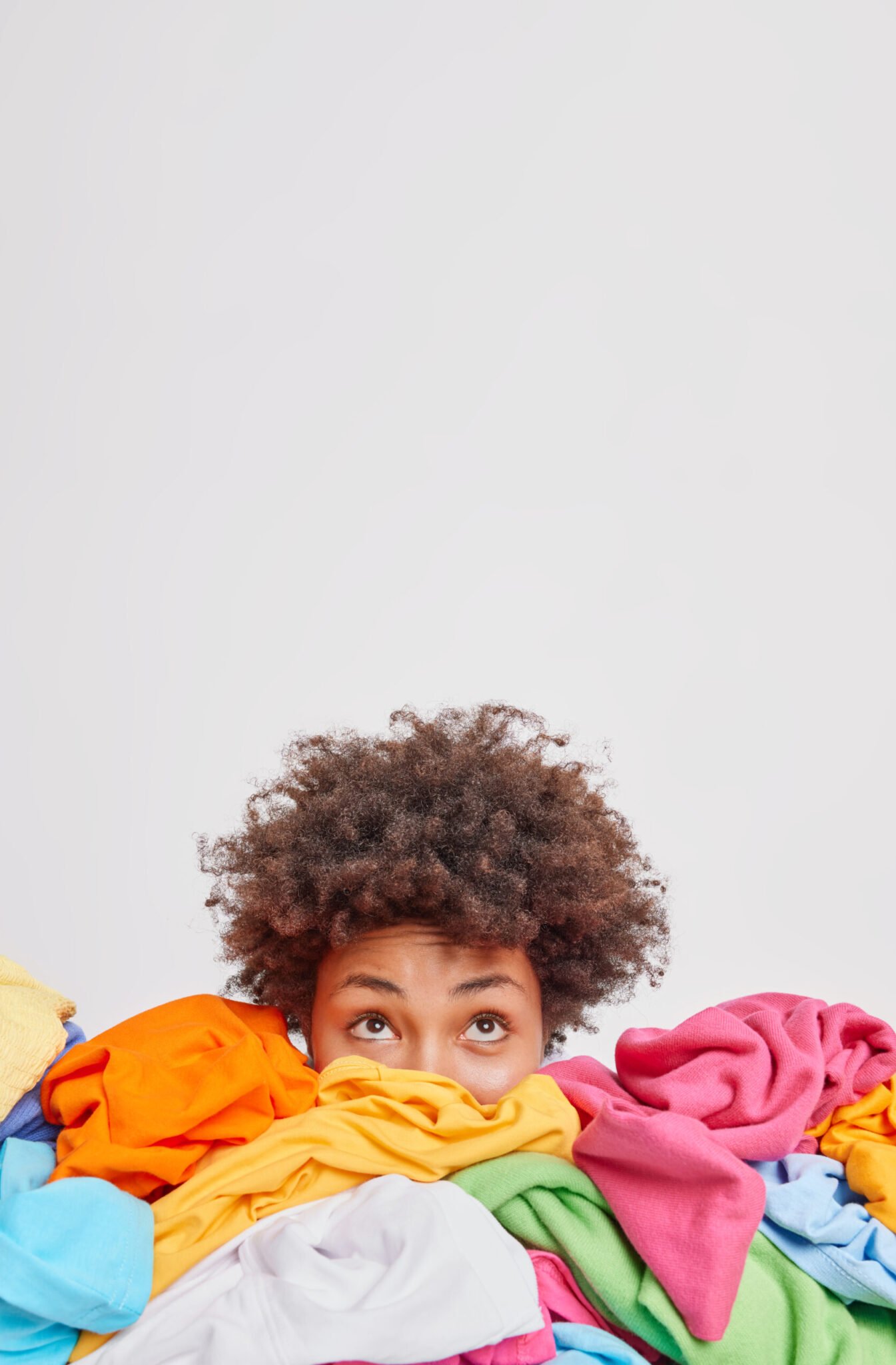 Young Afro American woman surrounded by different colorful clothes sorts out wardrobe focused above isolated over white background blank space for your advertising content. Nothing to wear concept